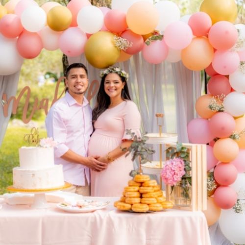 couple holding pregnant belly under a pink and gold balloon arch, standing in front of a cake