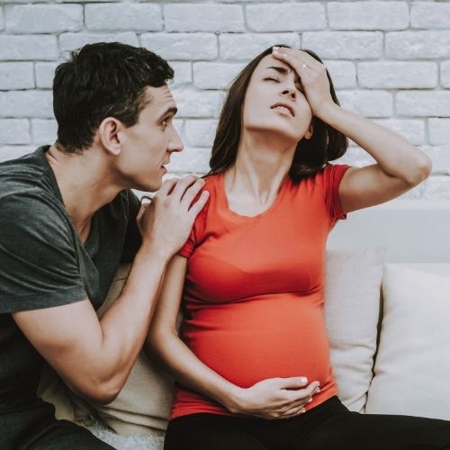 pregnant woman holding head and belly with partner leaning on shoulder