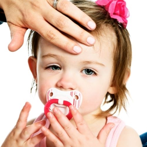 sick toddler girl sucking on pacifier with an adult hand against forehead
