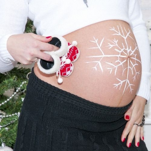 pregnant woman with snowflake painted on belly holding santa baby booties