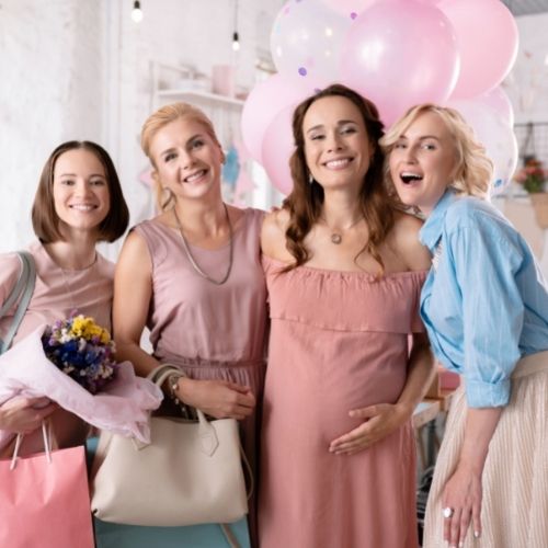 four women standing and smiling towards the camera. one woman holds her pregnant belly and there are pink balloons in the background of a baby shower