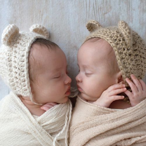 twins in neutral colors facing each other and sleeping while swaddled with crocheted bear hats on