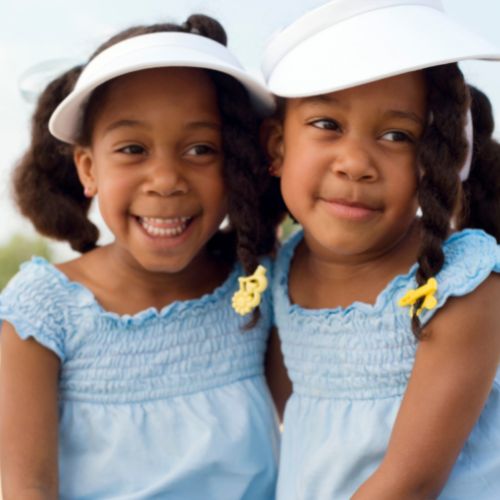 Two young girls wearing visors and dresses leaning against each other and smiling