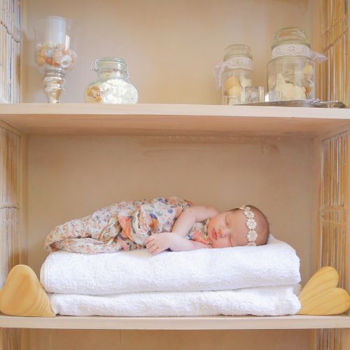 baby girl laying sideways on stack of two white towels on a bathroom shelf