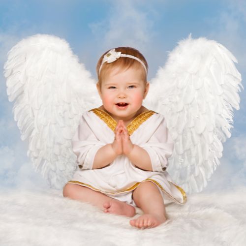 baby girl wearing a white dress with white angel wings over blue sky background with prayer hands and smiling