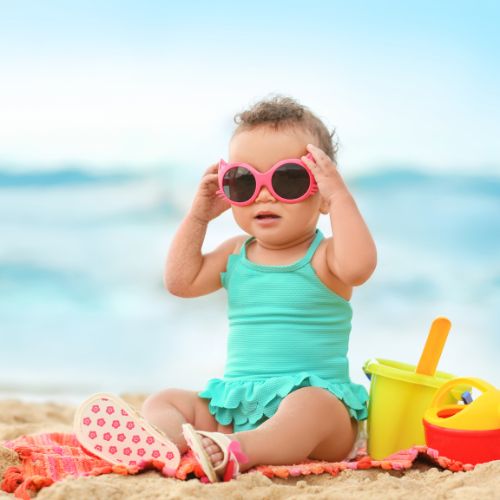 baby girl sitting up on beach with ocean in background wearing a bathing suit sitting near sand castle toys and wearing sunglasses