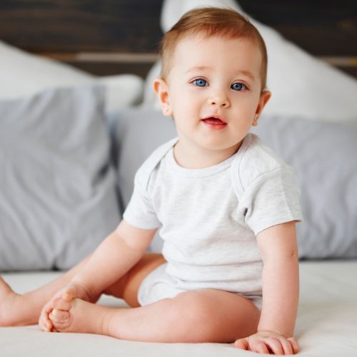 toddler child wearing white onesie sitting on bed looking at camera with gray pillows in the background