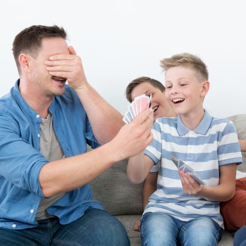 dad smiling and covering his eyes holding cards while two boys smiling pull at a card