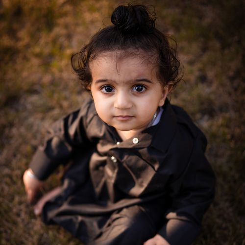 baby girl with dark curly hair sitting on grass and looking upwards towards camera wearing a black dress