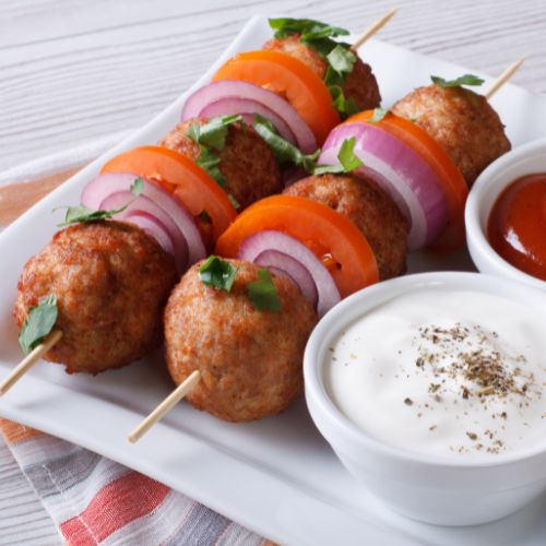 two skewers with meatballs, tomato slices and red onions on skewer. Skewers are on a white dish with two small bowls filled with one white sauce and one red sauce