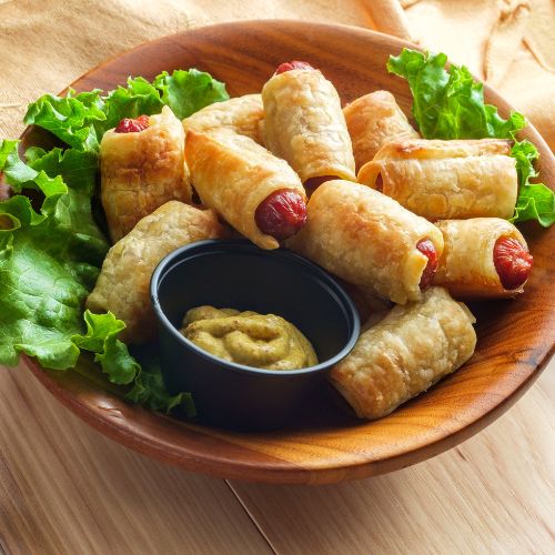 a small brown bowl with a lettuce garnish and a stack of crescent roll wrapped mini hotdogs with a small dish of dipping sauce