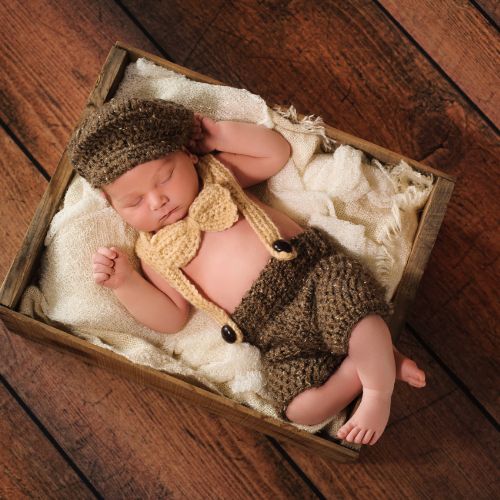 newborn baby boy wearing knitted brown pants with khaki colored knitted suspenders and a bow tie laying in a container on top of cream colored plush blankets wearing a brown hat on top of a wooden floor