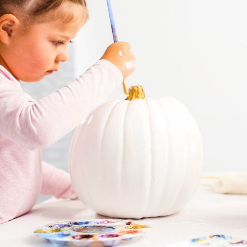 a toddler girl painting a pumpkin white with a paint brush and a focused look on her face