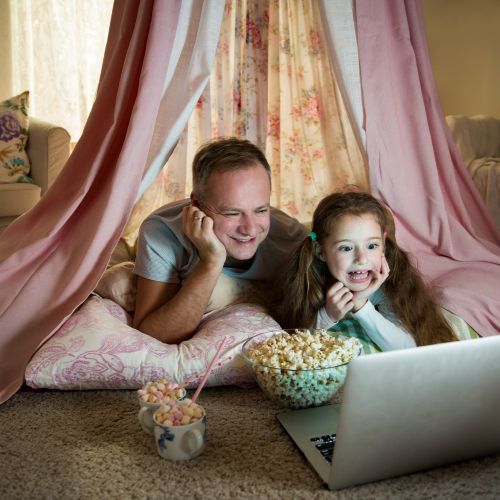 dad and daughter watching a movie on a laptop, smiling and laying on their bellies inside of an indoor play tent