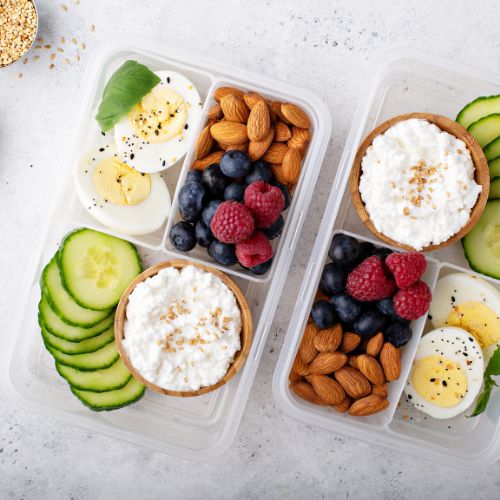 two snack boxes filled with cucumbers, cottage cheese, almonds, hard boiled eggs and berries